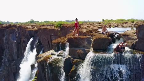 Aerial-Of-A-Man-And-Woman-Sitting-In-Devil'S-Pool-Waterfall-At-Victoria-Falls-Zambia