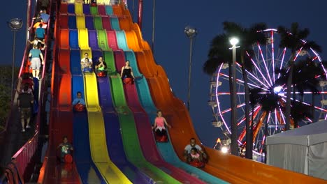 Los-Niños-Montan-Un-Gran-Tobogán-Colorido-Junto-A-Una-Noria-Por-La-Noche