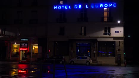 Ein-Hotelschild-Spiegelt-Sich-In-Den-Verregneten-Straßen-Von-Paris-Oder-Frankreich-Mit-Einem-Licht-Im-Obergeschoss-Wider