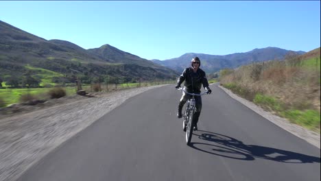 Un-Hombre-Con-Chaqueta-De-Cuero-Conduce-Una-Bicicleta-Motorizada-A-Lo-Largo-De-Una-Carretera-Rural-1