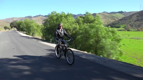Un-Hombre-Con-Chaqueta-De-Cuero-Conduce-Una-Bicicleta-Motorizada-A-Lo-Largo-De-Una-Carretera-Rural-4