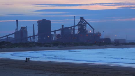 A-power-plant-at-dusk-along-a-beach-in-England-with-people-waking-background
