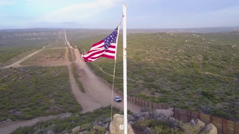 Die-Amerikanische-Flagge-Weht-über-Die-Us-mexiko-grenzmauer-In-Der-Kalifornischen-Wüste,-Während-Unten-Ein-Grenzpatrouillenfahrzeug-Vorbeifährt