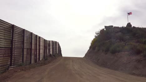 Eine-Leere-Straße-Entlang-Der-Mexikanischen-Grenze-Der-USA-Mit-Einer-Amerikanischen-Flagge-In-Der-Ferne
