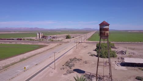 A-high-angle-aerial-over-a-lonely-abandoned-road-through-a-rural-area-past-water-tower-foreground