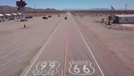 Antena-Drone-Sobre-Una-Carretera-Del-Desierto-Solitario-En-Arizona-Con-La-Ruta-66-Pintada-Sobre-El-Pavimento-Y-El-Coche-Pasando-Por-Debajo-De-1