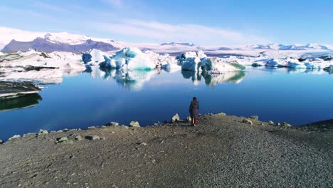 Aerial-of-a-woman-standing-along-the-shore-of-a-glacier-lagoon-in-the-Arctic-at-Jokulsarlon-glacier-lagoon-Iceland