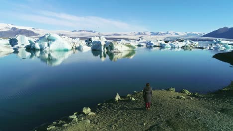 Antena-De-Una-Mujer-De-Pie-A-Lo-Largo-De-La-Orilla-De-Una-Laguna-Glaciar-En-El-Ártico-En-La-Laguna-Glaciar-Jokulsarlon-Islandia-3
