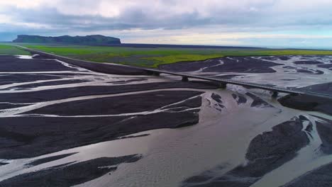 Antena-De-Un-Coche-A-Lo-Largo-De-Una-Carretera-Y-Un-Puente-Sobre-Un-Gran-Sistema-Fluvial-De-Inundación-Volcánica-Oscura-En-Islandia