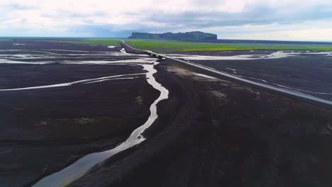 Antena-De-Un-Coche-Circulando-Rápido-A-Lo-Largo-De-Una-Carretera-Y-Un-Puente-Sobre-Un-Gran-Sistema-Fluvial-De-Inundación-Volcánica-Oscura-En-Islandia