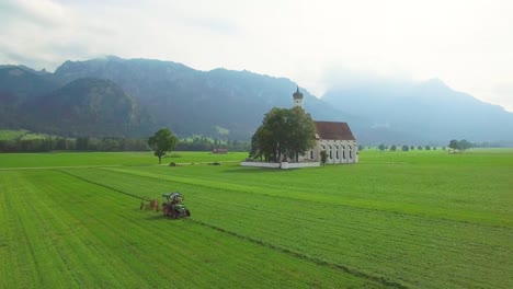 A-nice-aerial-over-a-traditional-German-Bavarian-church-with-a-tractor-plowing-fields-foreground