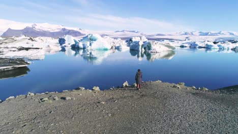 Antena-De-Una-Mujer-De-Pie-A-Lo-Largo-De-La-Orilla-De-Una-Laguna-Glaciar-En-El-Ártico-En-La-Laguna-Glaciar-Jokulsarlon-Islandia-1