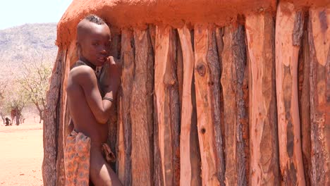 Un-Joven-Muchacho-Tribal-Himba-Africano-Se-Inclina-Contra-Su-Choza-De-Barro-Y-Madera-En-Una-Pequeña-Aldea-En-Namibia