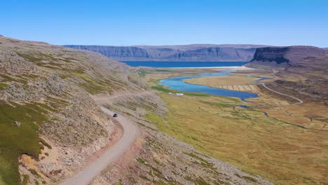 Beautiful-aerial-shot-of-a-car-traveling-on-a-dirt-road-with-the-Westfjords-and-fjords-of-Iceland-distant-