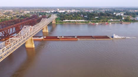Antena-De-Barcaza-Fluvial-Bajo-Tres-Puentes-De-Acero-Sobre-El-Río-Mississippi-Con-Fondo-De-Memphis,-Tennessee