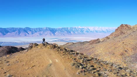 Drone-aerial-as-a-man-stands-on-the-edge-of-a-vast-open-view-of-the-Owens-Valley-desert-and-Owens-Lake-bed-in-the-Eastern-Sierras-of-California