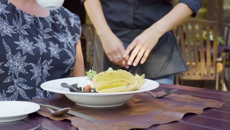 Una-Camarera-Entrega-El-Desayuno-A-Una-Mujer-En-Un-Restaurante-Al-Aire-Libre.