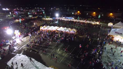 Antena-Sobre-Un-Carnaval-O-Feria-Celebrada-En-El-Estacionamiento-De-Un-Centro-Comercial-Con-Multitudes-Y-Puestos-Y-Atracciones