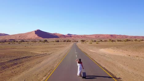 Aerial-over-a-woman-walking-with-a-suitcase-or-rolling-bag-on-a-lonely-road-in-the-Namib-Desert-in-Namibia-Africa
