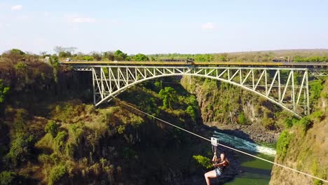 Aerial-of-a-woman-ziplining-across-a-canyon-with-Victoria-Falls-Bridge-background-Zimbabwe-and-Zambia-adventure-travel