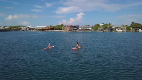 Aerial-over-two-women-rowing-paddleboards-towards-a-coastal-village-in-Honduras