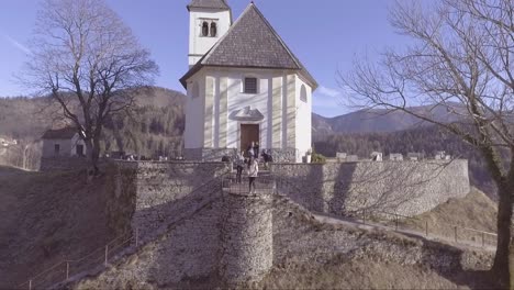 Vista-Aérea-of-a-small-white-church-in-the-mountains-of-Slovenia-and-woman-and-photographers