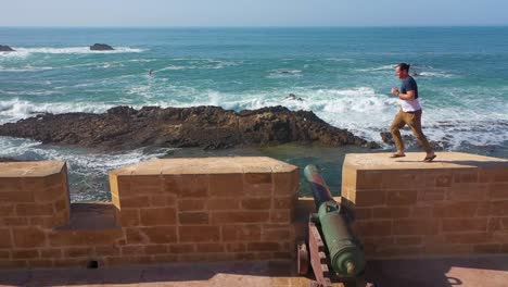 A-Man-Runs-Along-The-Ramparts-And-Jumps-Over-Cannons-In-The-Ancient-City-Of-Essaouira-Morocco