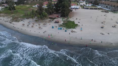 Antena-Sobre-Gente-Jugando-Al-Fútbol-En-La-Playa-De-Bakau,-Gambia,-África-Occidental