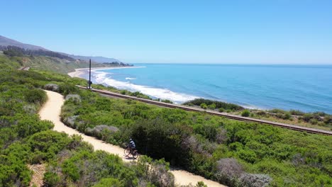 Aerial-Of-A-Man-Riding-His-Bicycle-Bike-With-Santa-Barbara-Mountains-And-Ocean-In-Background-Near-Carpinteria-Californa