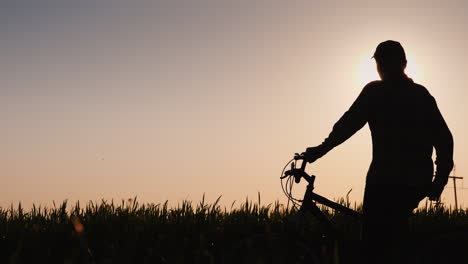 Die-Silhouette-Einer-Frau-Mit-Einem-Fahrrad,-Die-Auf-Einem-Feld-Steht-Und-Den-Sonnenuntergang-Anstarrt