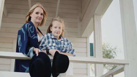 Mother-and-daughter-stand-on-the-porch-of-a-wooden-house-holding-hats-in-their-hands