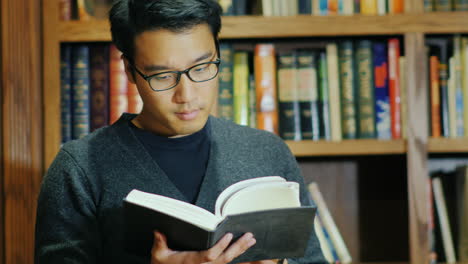 Apuesto-Hombre-Asiático-Con-Gafas-Leyendo-Un-Libro-En-La-Biblioteca