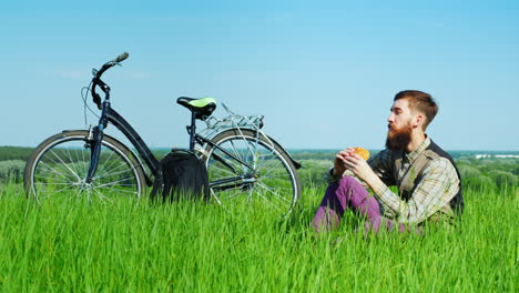 Young-Man-Eating-A-Hamburger-While-Sitting-On-A-Green-Meadow-Near-His-Bicycle