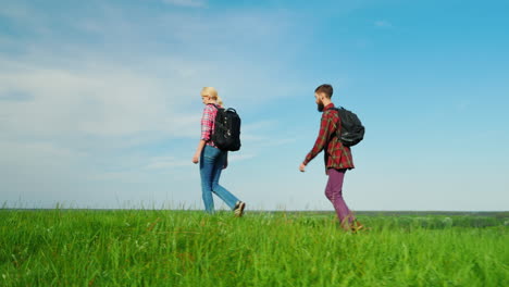 Two-Ecotourists-Are-Walking-Along-A-Green-Meadow-With-Backpacks-Behind-Them