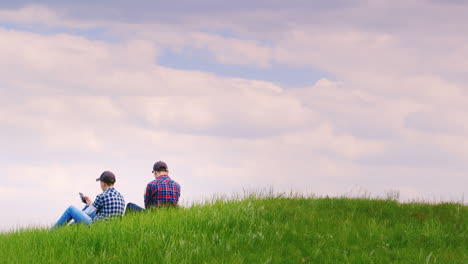 Two-Teenage-Boys-Are-Sitting-On-A-Green-Meadow-Using-Mobile-Phones-1