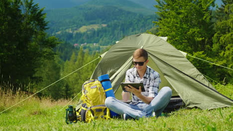 Ein-Junger-Männlicher-Tourist-Benutzt-Einen-Laptop-Beim-Camping-In-Der-Nähe-Eines-Zeltes