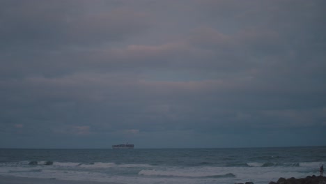 A-large-cargo-ship-sails-by-on-the-horizon-of-an-afternoon-beach-where-the-setting-sun-spills-light-colours-of-blue-and-pink-into-the-dangerous,-stormy-sky-as-well-as-the-dark-and-moody-water-below