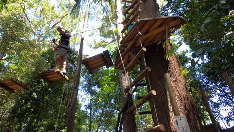 A-happy-Asian-man-steps-across-a-high-hanging-wooden-rope-bridge-in-a-treetop-adventure-park