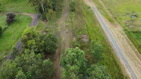 Overhead-drone-shot-down-tree-covered-dirt-path-as-two-cyclists-ride-towards-camera