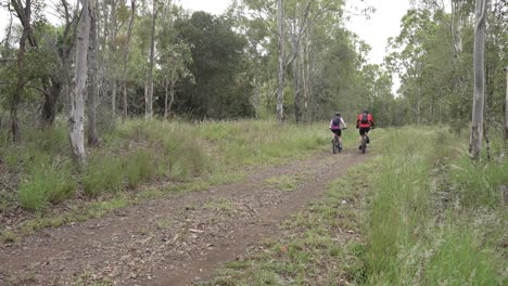 Two-cyclists-ride-past-camera-on-mountain-bikes,-down-a-dirt-road-in-Australian-bush