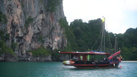 Beautiful-wooden-boat-is-anchored-behind-a-red-buoy-with-in-the-background-an-imposing-high-rock-mountain-of-Thai-nature-in-a-clear-blue-bay-in-Krabi