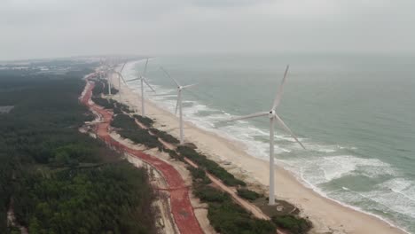 Array-of-onshore-horizontal-axis-wind-turbines-located-on-a-long-beach-coastline-with-high-ocean-swell-on-a-cloudy-day