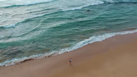 Girl-alone-on-a-beach-staring-at-beautiful-blue-ocean-water-angled-aerial-view