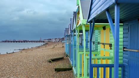 Stylish-and-smooth-dolly-shot-looking-along-a-line-of-colorful-traditional-English-beach-huts-on-a-shingle-beach-at-English-seafront-town-of-Herne-Bay-with-pier-in-the-background