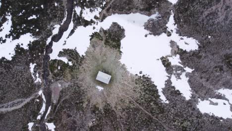 Aerial-drone-footage-slowly-descending-towards-a-remote-bothy-with-beautiful-patterns-in-the-grass,-snow-patches,-a-river-and-frosty-moorland-in-winter