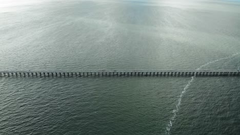 Push-and-fast-camera-tilt-towards-long,-wooden-pier-stretching-outwards-into-the-great-expanse-of-dark,-gloomy-blue-water-on-a-sunny-day