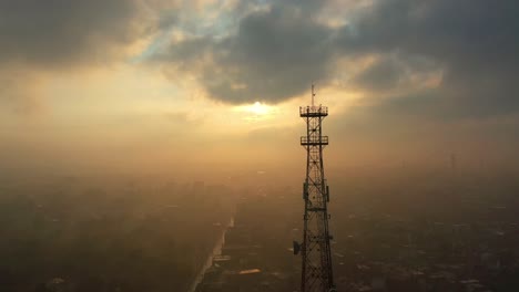 Vista-Aérea-De-La-Torre-De-Telecomunicaciones-Contra-La-Luz-Del-Atardecer-A-Través-Del-Aire-Brumoso-En-La-Distancia-Sobre-La-Ciudad-De-Moro-Sindh