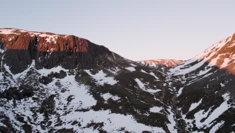 Aerial-drone-footage-panning-to-reveal-steep,-dramatic-mountains-and-cliffs-in-a-winter-landscape-with-snow,-boulders-and-rocks-at-sunrise-in-a-remote-part-of-the-Cairngorms-National-Park,-Scotland