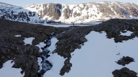Aerial-drone-footage-flying-away-from-two-people,-a-river-and-Loch-Etchachan-in-a-frozen-winter-landscape-of-snow-covered-mountains,-vertical-cliffs-of-steep-rock,-snow-and-ice