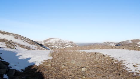 Aerial-drone-footage-reversing-over-a-rocky-cliff-with-clear-blue-skies-in-the-mountains-near-Ben-Macdui-in-the-Cairngorms-National-Park,-Scotland-as-sunshine-reflects-off-of-snow-and-boulders